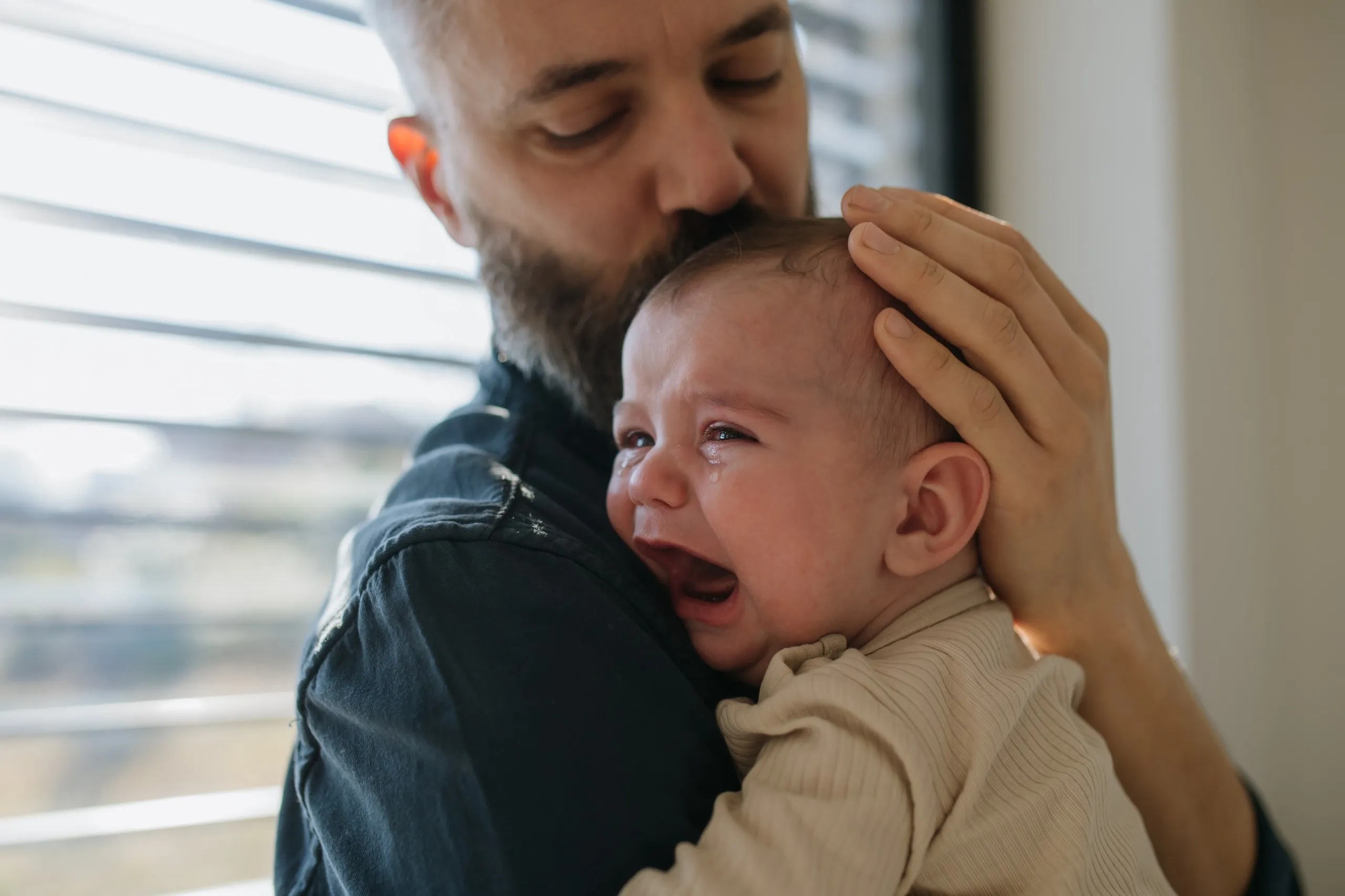 Padre abrazando y besando la cabeza de su bebé, que llora sobre su hombro junto a una ventana